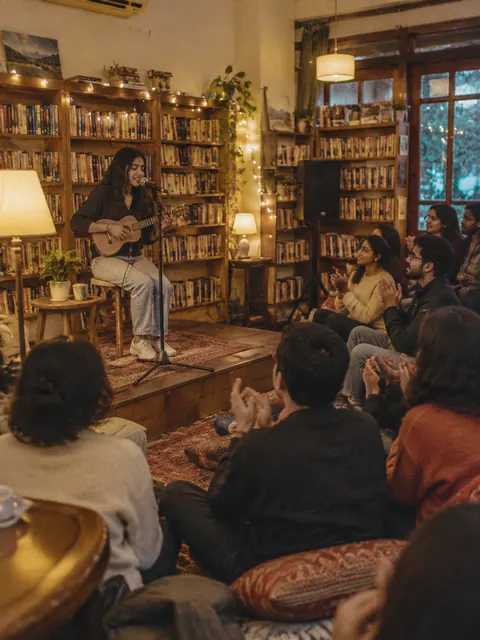 A woman singing with a ukulele at a small open mic in a bookshop cafe.