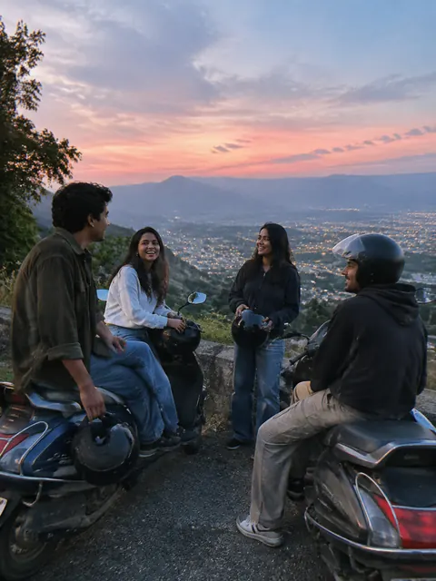 Four friends with two scooters at a hillside viewpoint at dusk, city lights below.